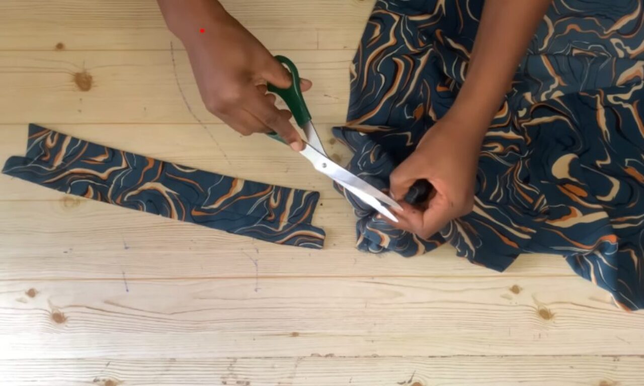 Hands of a person cutting a piece of patterned fabric with green-handled scissors on a light wooden table. The fabric has a swirling design in blue, orange, and cream colors, and a strip has already been cut and placed to the side.