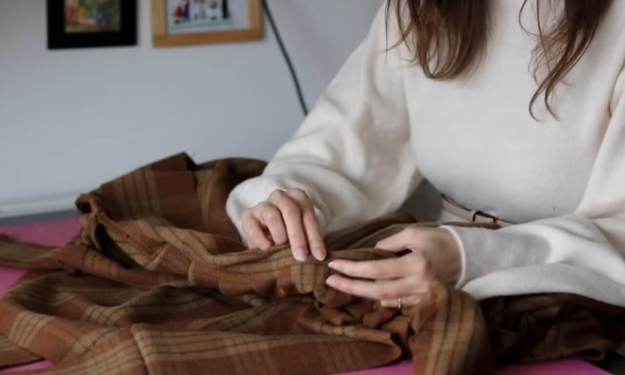 A person wearing a cream-colored sweater is sitting at a table, using their hands to sew or inspect a brown plaid fabric. Only the person's torso and hands are visible. There are framed pictures on the wall in the background.
