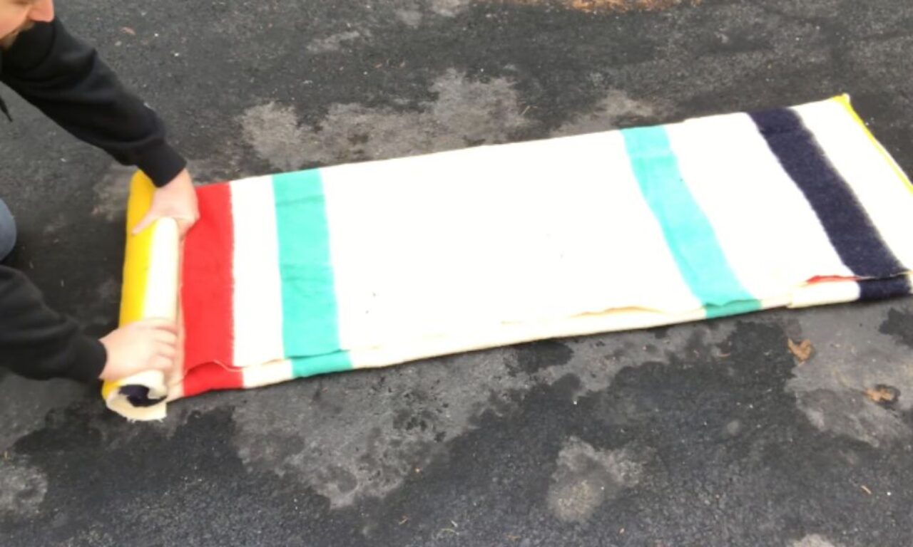 A person rolls up a white blanket with bold, colorful horizontal stripes on a dark paved surface. The stripes, from left to right, are yellow, red, turquoise, and navy blue.