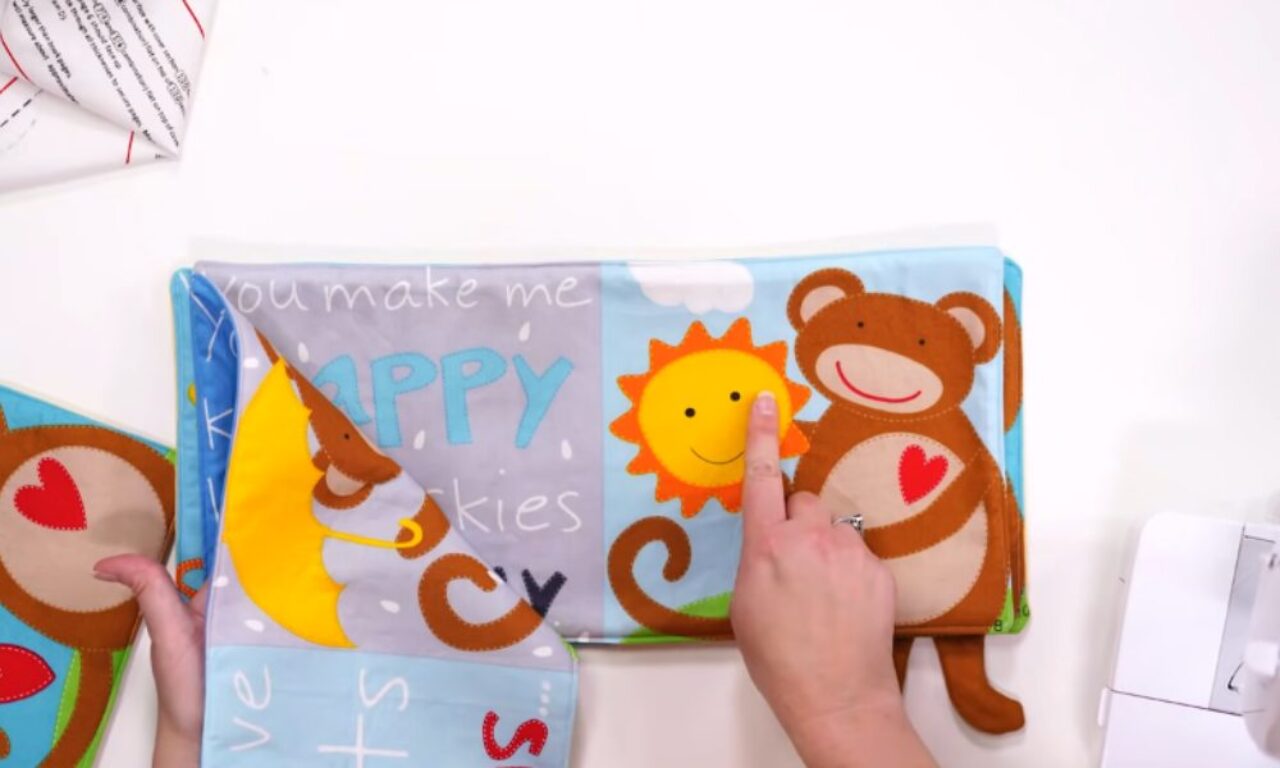 A person’s hands are holding and pointing to the pages of a colorful fabric children’s book. The open page shows a smiling cartoon monkey with a red heart on its chest and a cheerful sun with a face. The background is light blue with white clouds. Part of the left page, with the word "HAPPY" and a yellow crescent moon, is being lifted. A sewing machine is partially visible on the right side of the image.