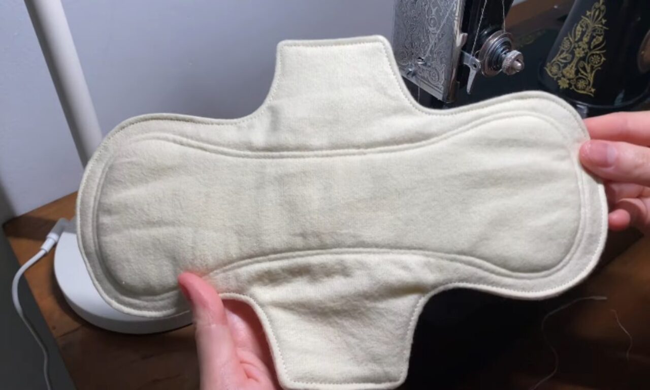 a handmade, off-white reusable cloth menstrual pad. The pad is held by a pair of hands, showcasing its shape and stitching. In the background, a vintage sewing machine is partially visible, suggesting the pad was recently made.