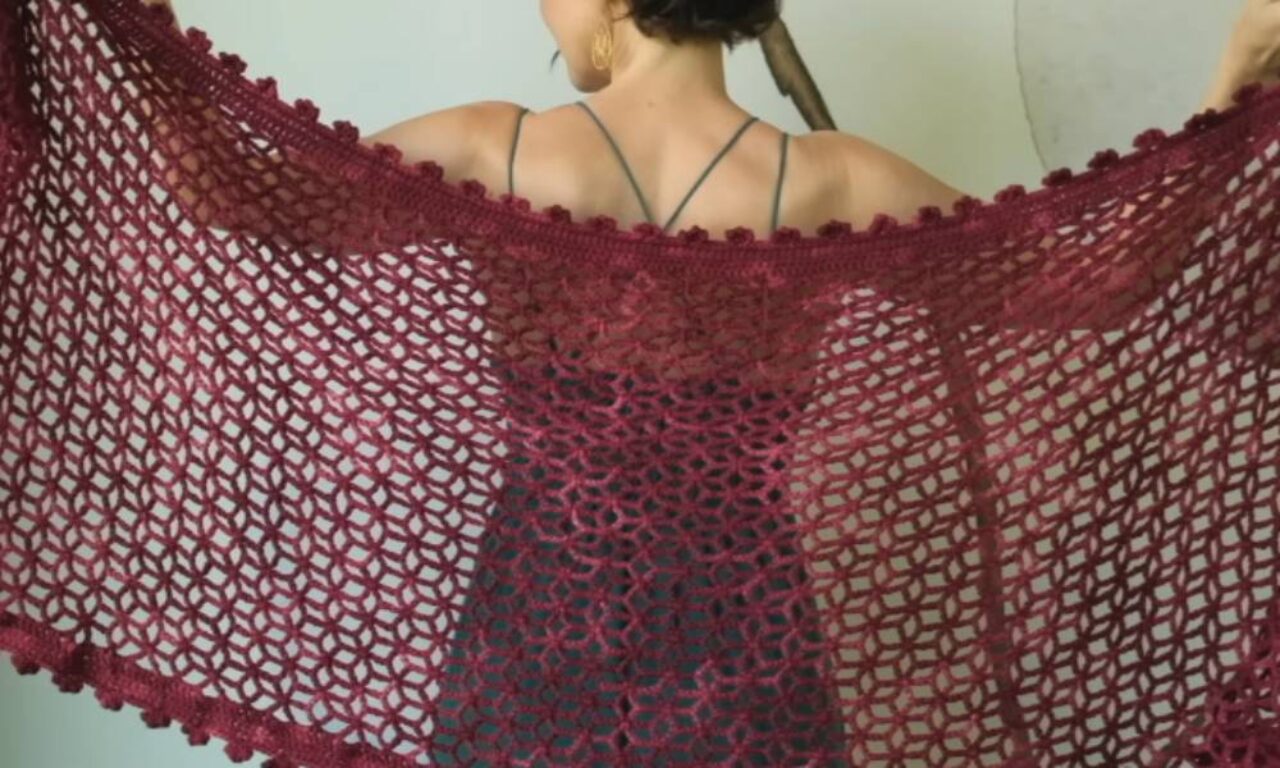 Back view of a person in a strappy dress holding their arms wide to display an oversized, burgundy, open-lattice crochet shawl with a scalloped border.