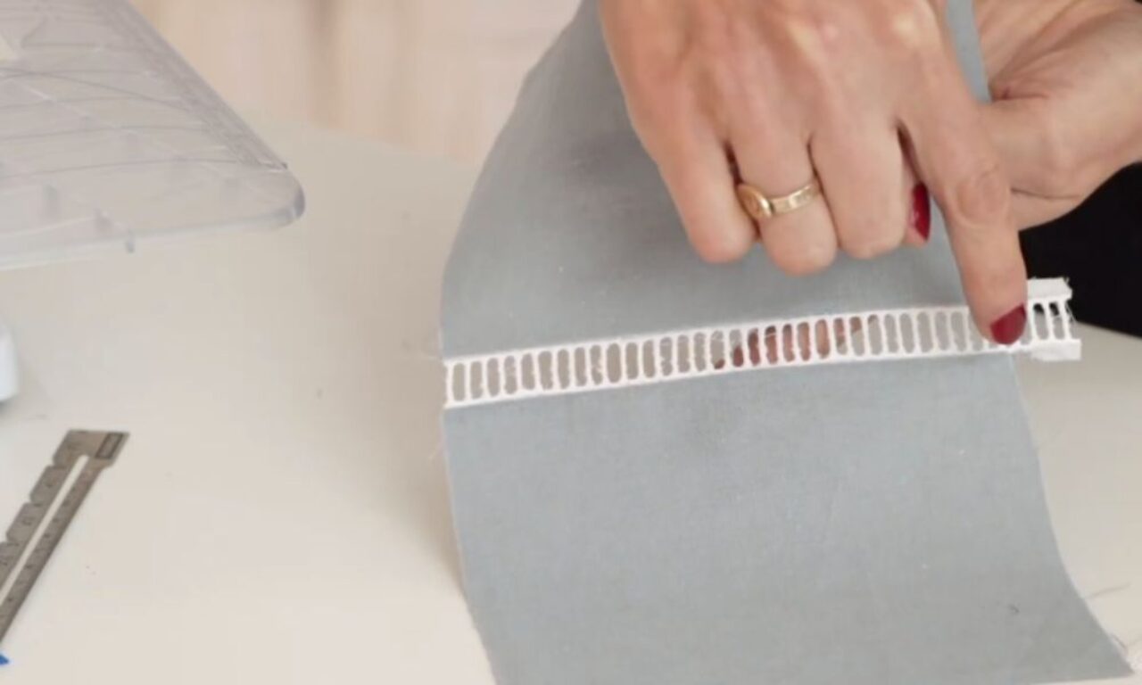 A hand points to a white perforated trim attached to light grey fabric.