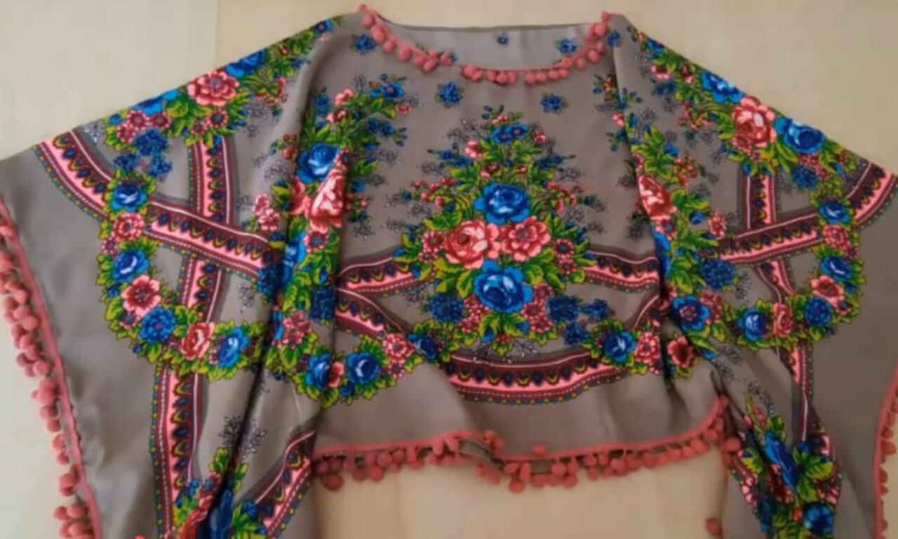 Flat lay of a poncho-style garment made from a taupe fabric with a traditional, brightly coloured floral folk-art print (pinks and blues). Dusty-pink pom-pom trim edges the round neck and the entire draped hem.