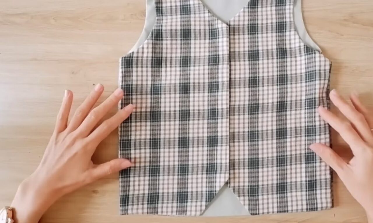 A person is smoothing out a plaid vest pattern on a light wooden surface, with both hands gently pressing the fabric to remove wrinkles. The vest is made of a white, black, and gray plaid fabric and has a gray lining visible around the armholes and neckline.