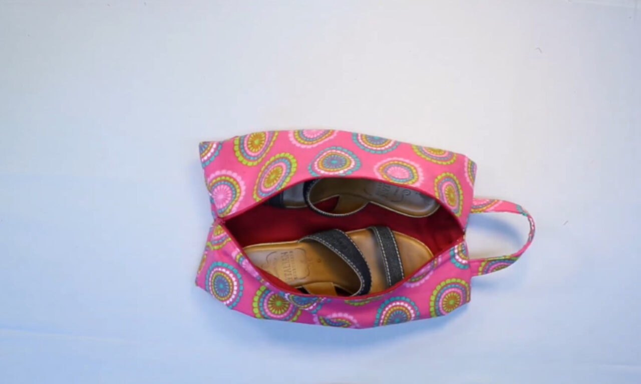 A bright pink fabric shoe bag, adorned with a colorful circular medallion pattern, lies open on a plain light background. Inside, nestled against the red lining, are two pairs of women's sandals—one with dark fabric straps and another with lighter straps. The bag has a matching fabric handle on the right side.