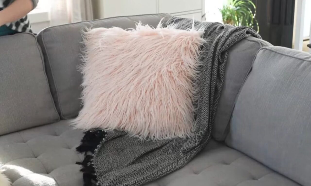 A gray sofa with tutufted seat cushions is adorned with a fluffy, light pink faux fur pillow and a dark gray throw blanket featuring a geometric pattern and tassels. Bright sunlight illuminates the front left side of the couch.