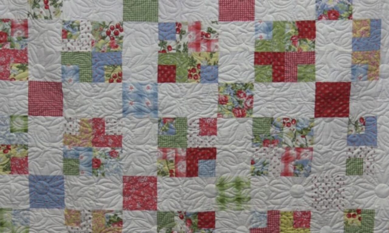 A close-up view of a quilt featuring a patchwork design with small squares and L-shaped patterns in shades of red, green, blue, and floral prints, all set against a white background with intricate swirling quilted stitching.