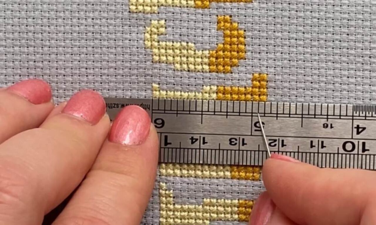 A close-up of cross-stitch embroidery in progress on gray aida cloth. The pattern features yellow and gold thread forming what appears to be a decorative motif. A metal ruler is being held against the fabric to measure the work, with fingers visible holding both the ruler and fabric. The stitches show the characteristic X pattern of cross-stitch technique.