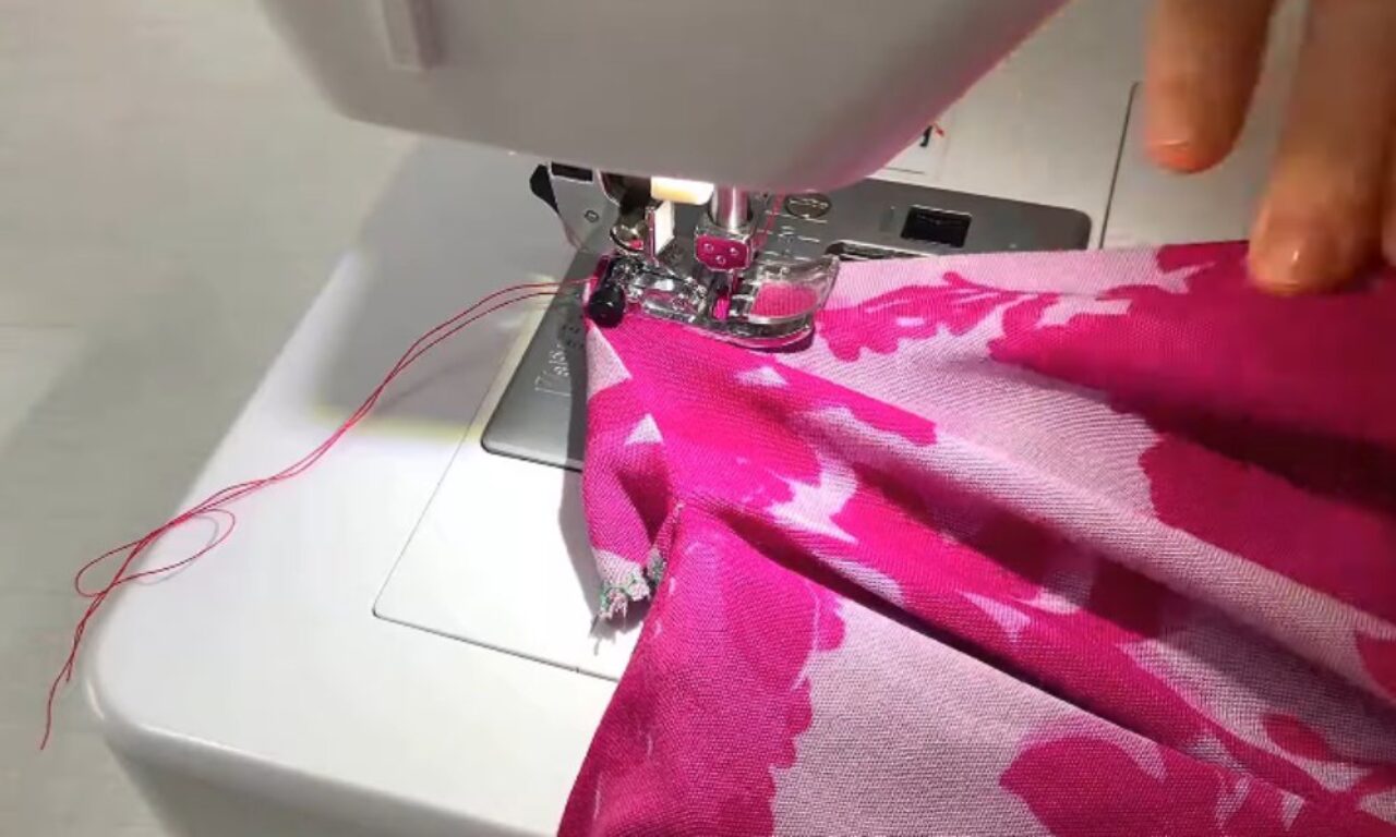 A close-up of a sewing machine in use, stitching pink and white patterned fabric. A person's hand is guiding the fabric under the sewing machine needle, and pink thread is visible trailing to the left.