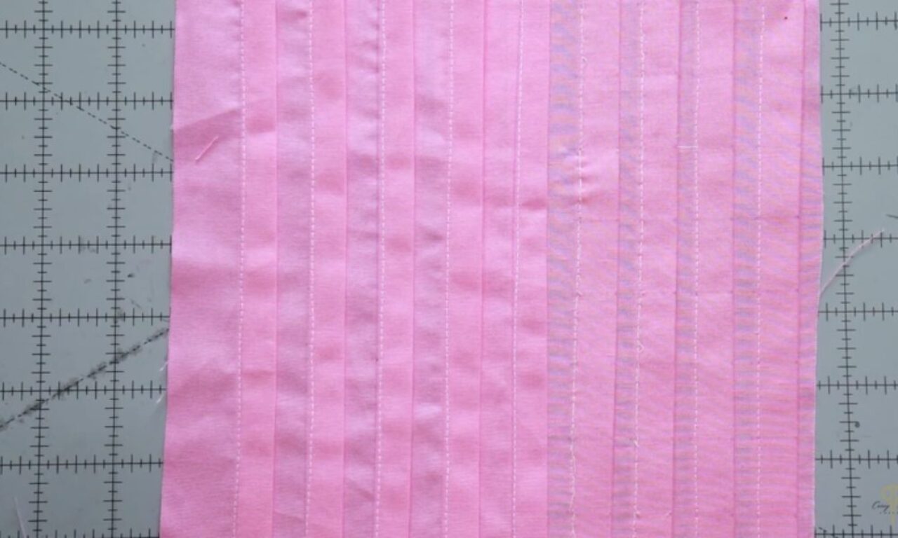 A piece of pink fabric placed on a gray cutting mat with measurement markings. The fabric has vertical lines of stitching, demonstrating different sewing techniques or stitch tensions.
