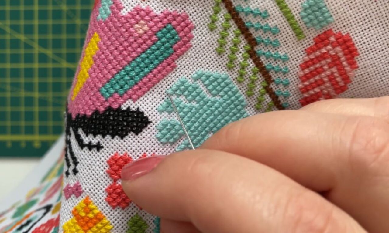 A close-up of a person’s hand guiding a needle through a colorful cross-stitch design with pink, teal, yellow, red, and black shapes, set against a green cutting mat in the background.