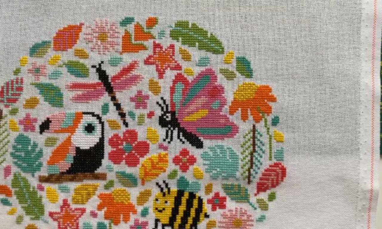 Colorful cross-stitch embroidery featuring a circular jungle-themed design. Inside the circle are vibrant images of a toucan, butterfly, dragonfly, bee, and various flowers and leaves in bright shades of pink, orange, yellow, green, and turquoise. The background fabric is a light neutral color, and the edge of the fabric is visible on the right.
