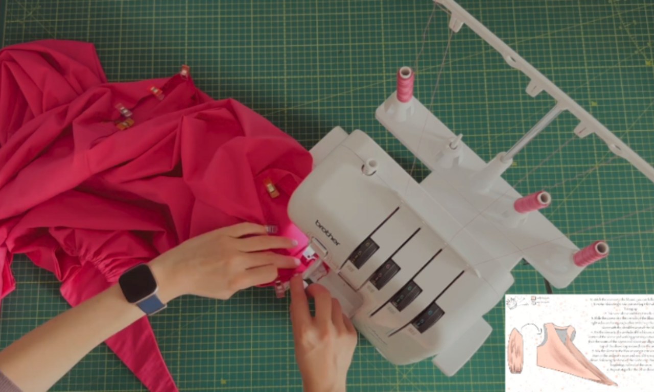 A person is using a white Brother overlock sewing machine to sew a piece of bright red fabric. The fabric is held in place with yellow safety pins, and the person's hands are guiding it through the machine. The workspace features a green cutting mat with grid lines, and there is an instruction sheet or diagram visible on the right side of the image. The scene suggests a sewing or crafting project in progress.