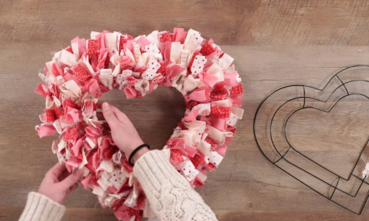A person's hands tie pieces of pink, white, and red patterned fabric onto a heart-shaped wire frame to create a fluffy wreath. A larger, empty heart-shaped wire frame is beside it on a wooden surface.