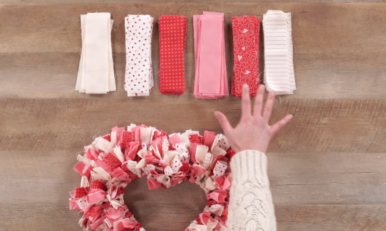 A person's hand, wearing a white knitted sleeve, is pointing to a colorful fabric wreath made from various shades of red, pink, and white ribbons. Above the wreath, there are six rolls of fabric in different colors and patterns, including solid white, white with red polka dots, solid red, solid pink, red with white heart patterns, and white with red stripes.