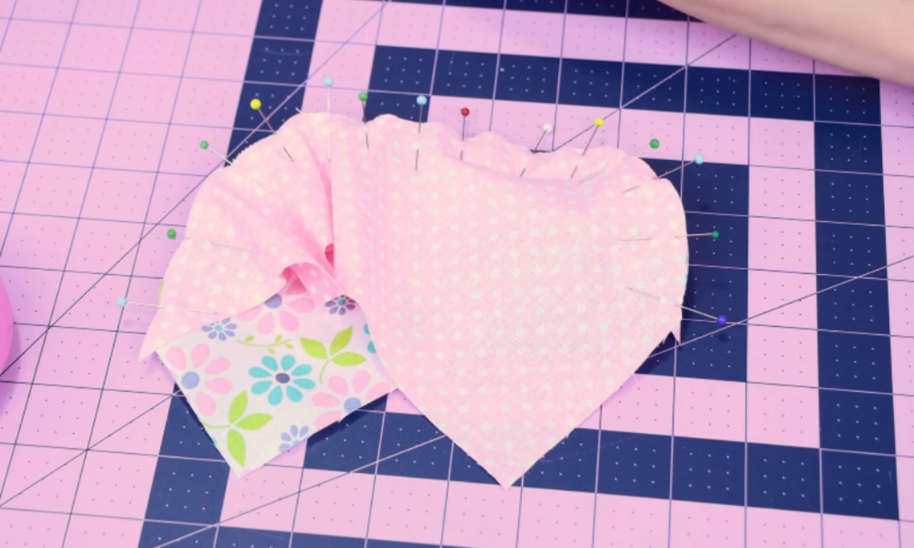 The image shows a piece of pink fabric with a floral pattern, cut into the shape of a heart, placed on a cutting mat with a grid pattern. The heart-shaped fabric is pinned in place with several colorful sewing pins. There is also a smaller piece of fabric with a floral design in pastel colors, partially visible beneath the heart-shaped fabric. The cutting mat has a grid layout with alternating pink and blue squares, which is commonly used for precise cutting and sewing projects.