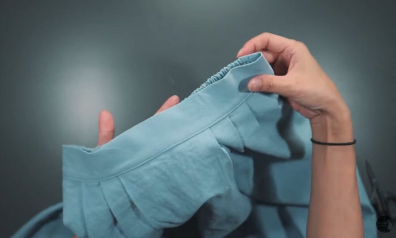 A pair of hands holding and examining a light blue fabric garment. The hands are gently manipulating what appears to be a shirt or blouse with pleated details and an elastic or gathered edge. The person is wearing a thin black bracelet on one wrist. The background is a soft, muted gray.