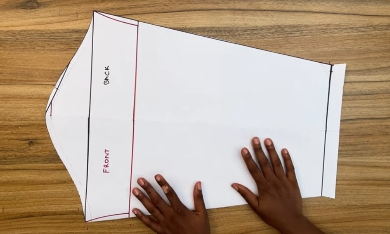 Two hands are placed on a white paper sewing pattern laid flat on a wooden surface. The pattern is labeled with "FRONT" and "BACK" sections, with lines drawn in black and red ink to outline the shape. The paper appears to be folded or creased in some areas.
