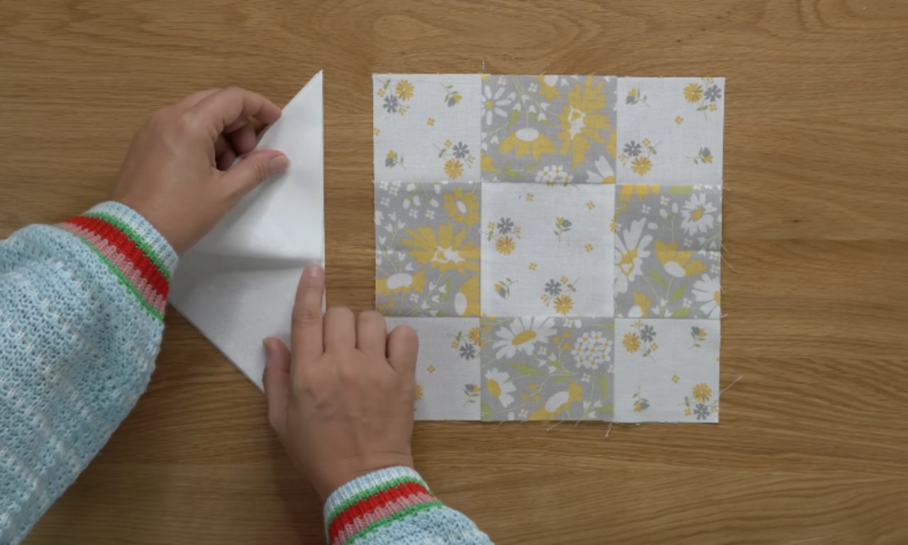 Hands holding a piece of paper over a completed sampler quilt block with various floral and geometric patterns. The quilt block is displayed on a wooden surface. The image is from a YouTube video titled 'Oh, Happy Day! Sampler Quilt Sew Along WEEK 4 [Best Loved Block]' by Coriander Quilts.