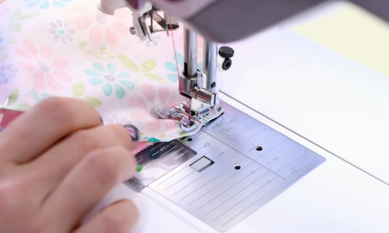 Close-up of a person's hand guiding a piece of floral-patterned fabric under the presser foot of a sewing machine, with the needle and thread visible as they stitch the fabric.
