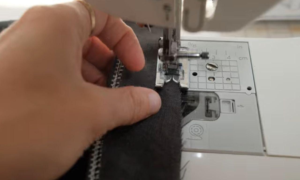 A close-up shows a person's hand guiding dark fabric under the presser foot of a white sewing machine. White stitching is visible along the left edge of the fabric, and the machine's metal throat plate with measurement markings is in the foreground.