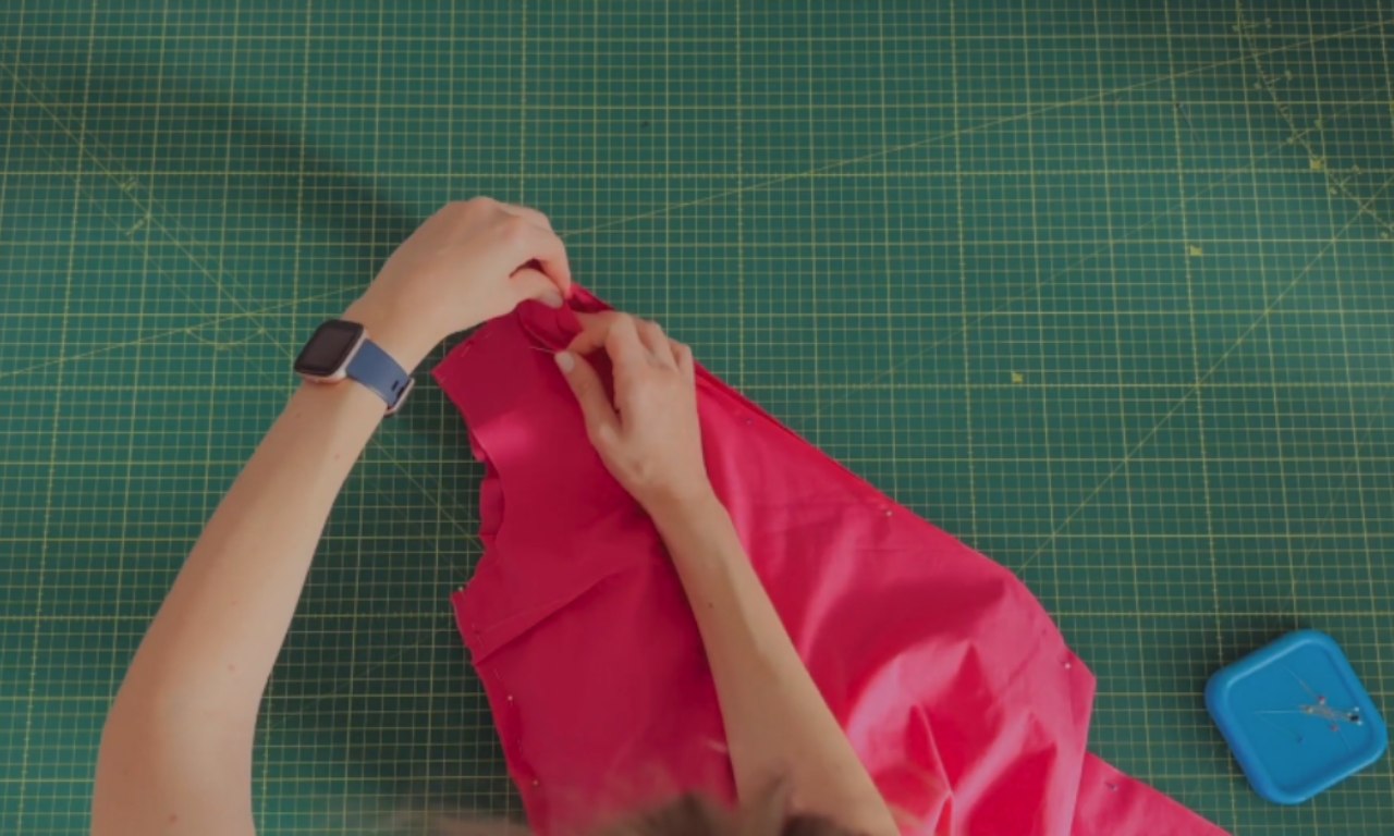 Top-down view of a person’s hands pinning a bright pink garment piece on a green gridded cutting mat; a blue pin cushion sits nearby and the person wears a smartwatch with a blue band.