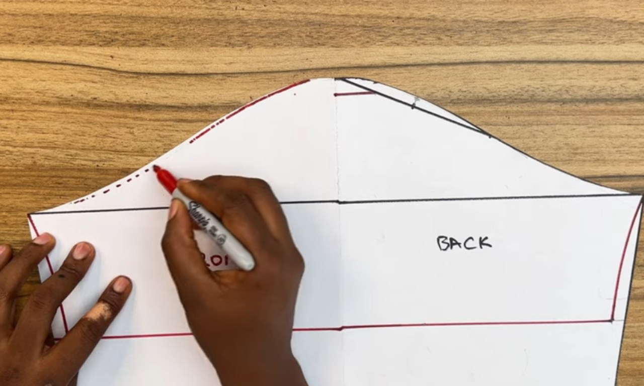 A person's hand uses a red marker to draw a curved line on a white paper sewing pattern. The pattern, laid out on a wooden surface, shows existing black lines, including a dashed line being traced, and the words 'FRONT' (partially visible) and 'BACK' on different sections, indicating it's a sleeve pattern piece.