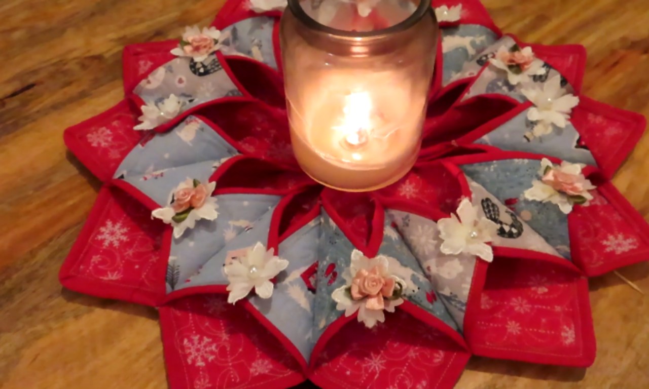 a beautifully crafted fabric wreath, ideal for seasonal decoration. The wreath features a red and white color scheme with floral patterns and is adorned with small white and pink fabric flowers. At the center of the wreath, there is a lit candle placed inside a glass jar, adding a warm and cozy ambiance.