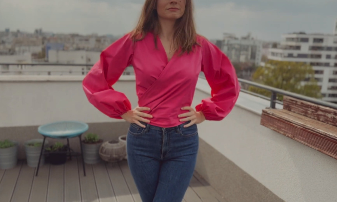 A woman wearing a vibrant pink wrap blouse with voluminous, puffy sleeves and dark blue jeans stands with her hands on her hips on a rooftop. In the blurred background, city buildings are visible under a cloudy sky.