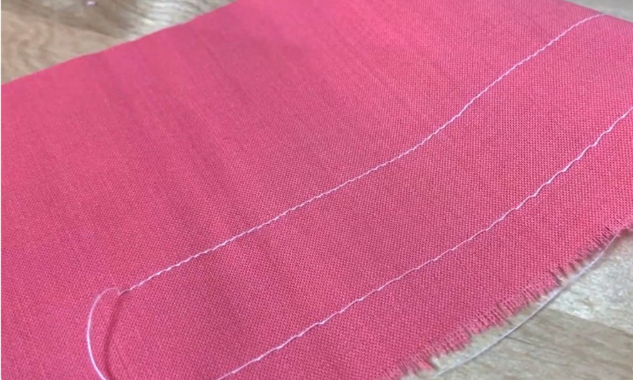 Pink fabric with white basting stitches outlining a curved shape, placed on a wooden surface.
