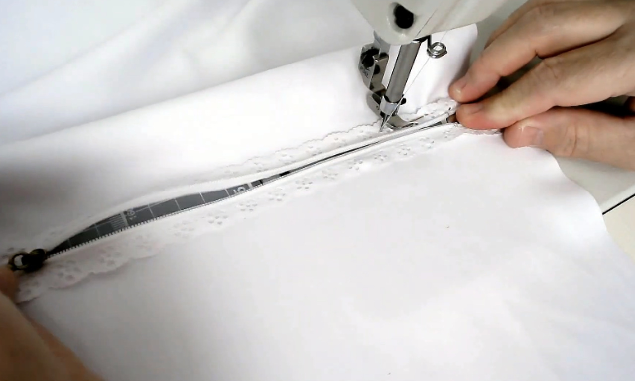 a white zipper being carefully guided under the presser foot of a sewing machine. The zipper is being attached to white fabric trimmed with delicate lace. Hands are visible guiding the fabric.
