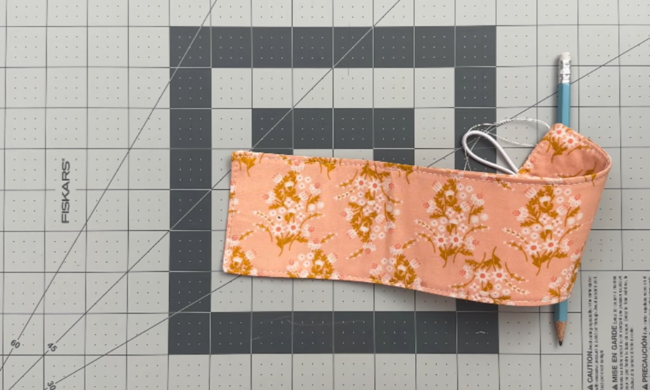 A peach-colored fabric bookmark or pen holder with a repeating pattern of white and yellow-gold flowers and green stems. The rectangular fabric item is folded and has a white elastic loop extending from one end. A light blue pencil is tucked into the opposite end. The item is resting on a gray self-healing cutting mat with a grid pattern.