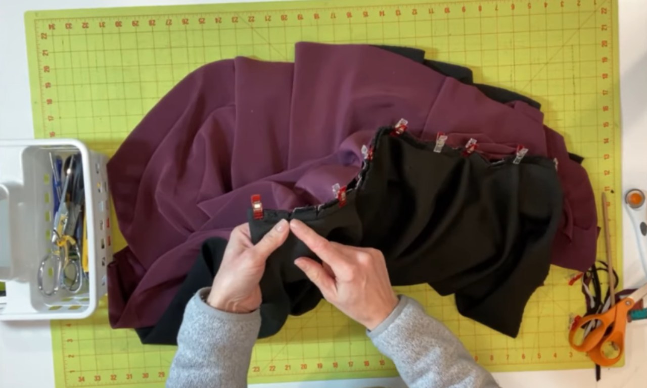 hands manipulating two pieces of fabric – black on top, maroon underneath – that are joined by red sewing clips along a seam. The hands point to the clipped seam. The fabrics rest on a green grid cutting mat, with a white caddy of sewing tools on the left and scissors and a rotary cutter on the right.