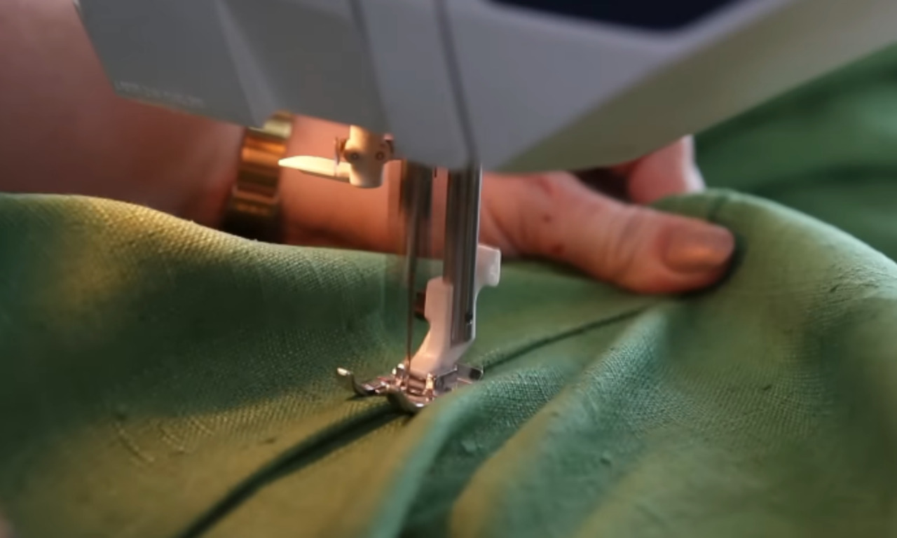 A close-up, low-angle view of a sewing machine's needle and presser foot actively stitching a seam on bright green, textured fabric. A person's hand is visible on the right, guiding the fabric.