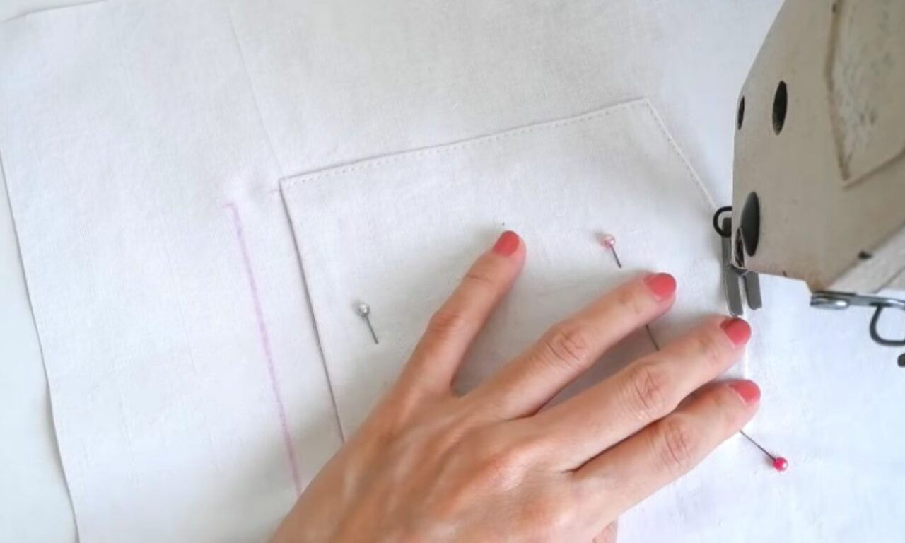 A person is sewing a square pocket onto a piece of white fabric using a sewing machine. The person's hand is guiding the fabric, and there are pins holding it in place. The person's fingernails are painted pink.