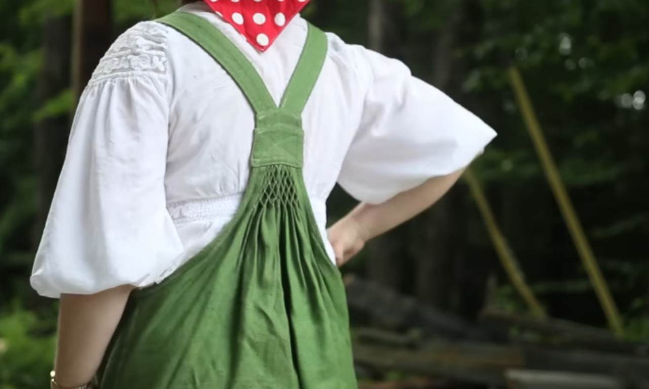 A back view of a person wearing a white blouse with puffy sleeves and a green pinafore dress. The straps of the dress cross and connect to an intricately smocked panel with a diamond pattern above a gathered waist.