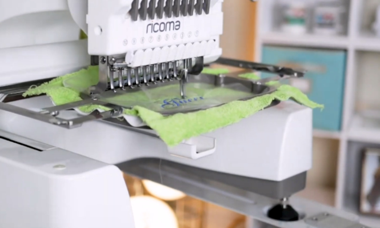 an embroidery machine in action. The machine, branded "Ricoma," is embroidering a design onto a piece of light green fabric. The fabric is held in place by a hoop, and the needles are actively stitching the pattern. The background is slightly blurred, featuring shelves with various storage boxes and containers, indicating a well-organized workspace. The focus is on the embroidery process, highlighting the precision and detail involved in creating embroidered designs.