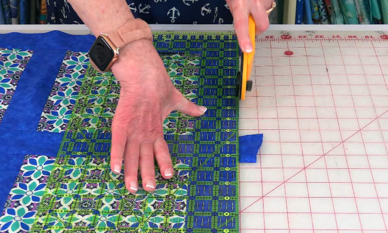 A person uses a yellow rotary cutter and a clear quilting ruler to trim the edge of a blue and green floral quilt top on a gridded cutting mat.