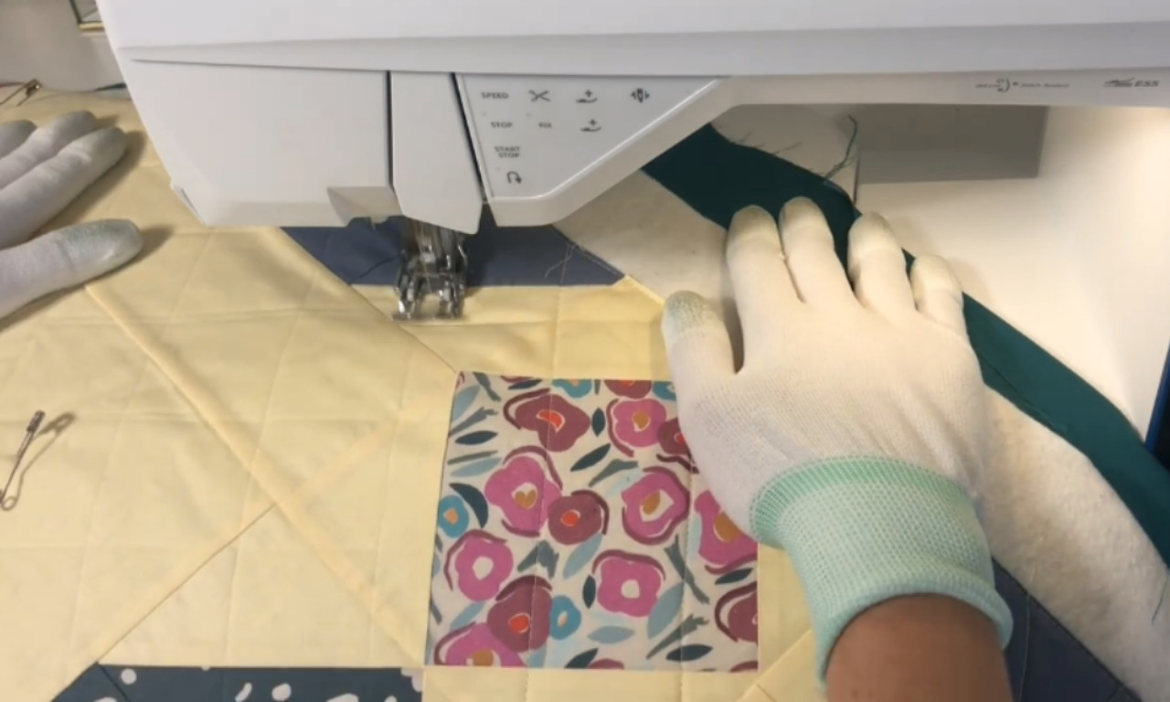 a person's hands, wearing white quilting gloves, guiding a colorful quilt under the needle of a modern white sewing machine. The quilt features light yellow fabric with diagonal quilting lines, a central block with a pink, purple, and blue floral pattern, and a dark teal fabric strip being attached along the edge as binding. A safety pin is visible on the yellow fabric to the left.