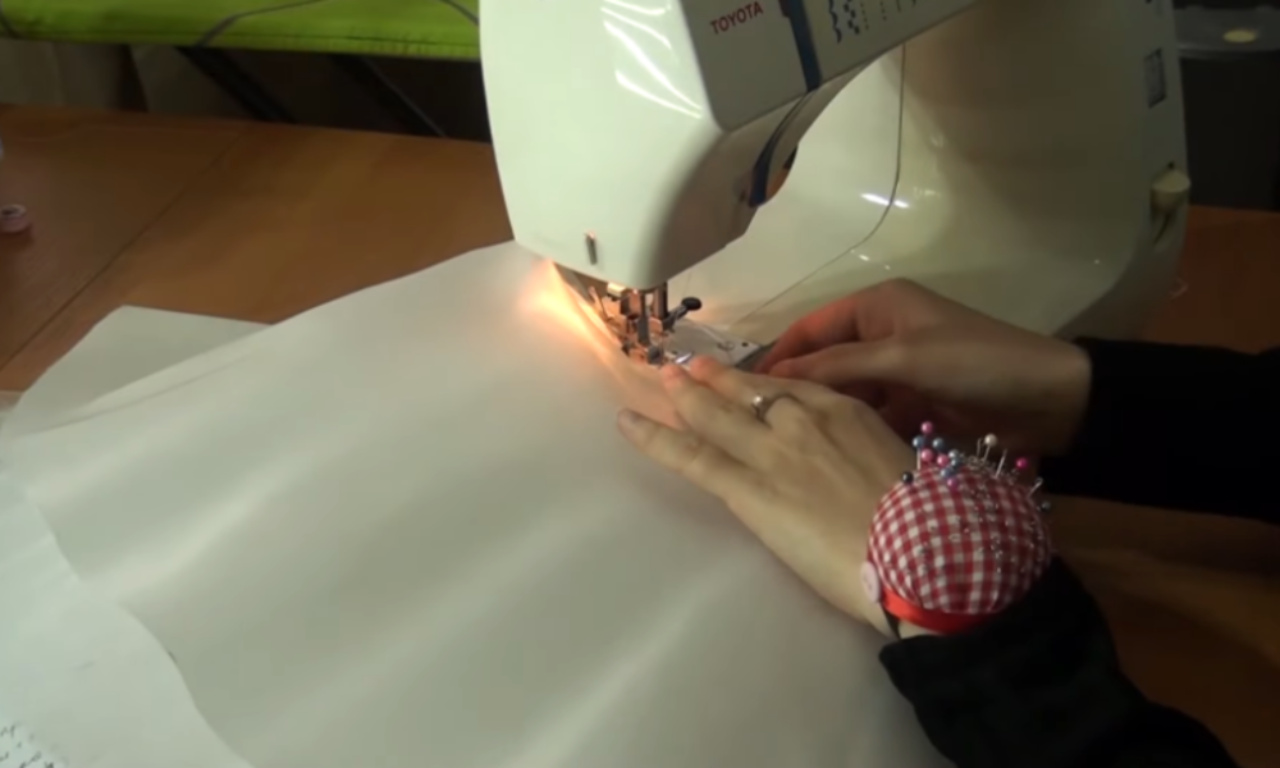 a person's hands guiding white fabric through a white Toyota sewing machine. The machine's needle area is illuminated by a bright light. A red and white checkered pin cushion, filled with colorful pins, is worn on the person's wrist.