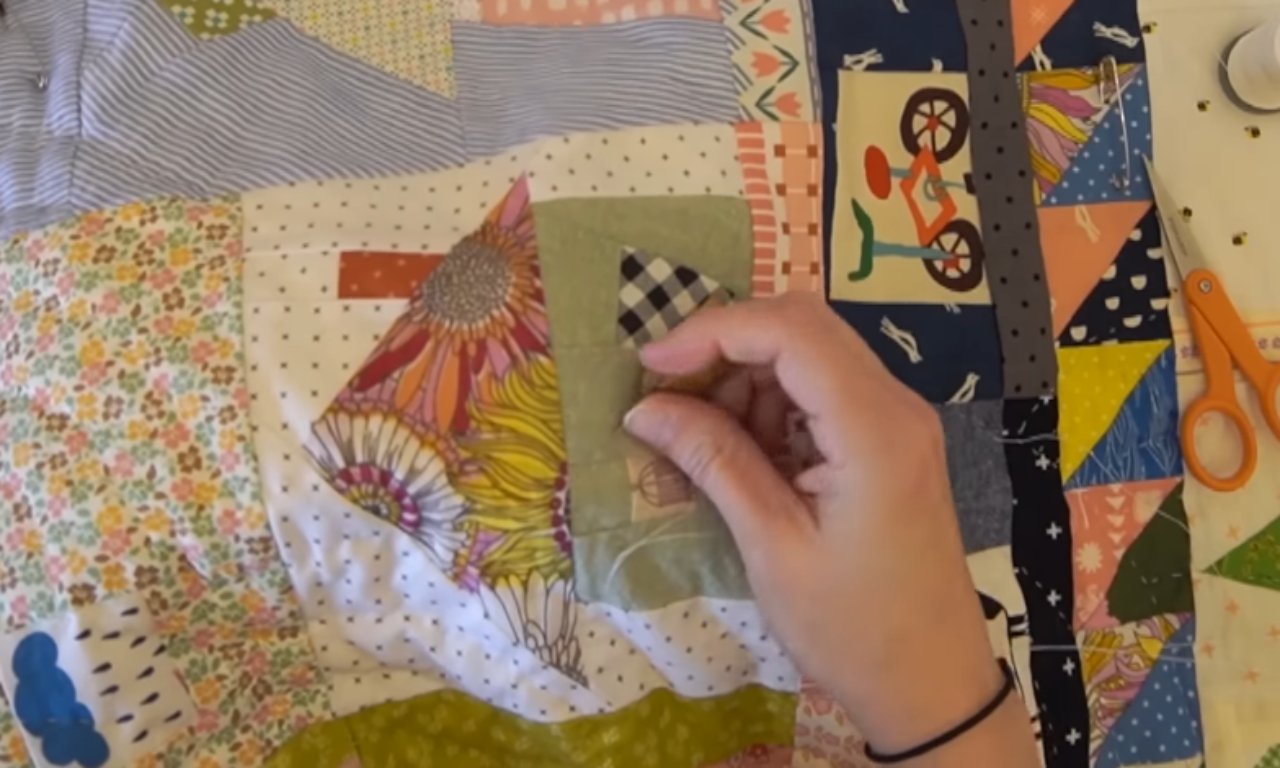 A close-up of a person's hand sewing a patch onto a colorful, patterned quilt. The quilt features various fabric designs, including floral, checkered, and geometric patterns. Scissors and a spool of thread are visible on the right side of the image.