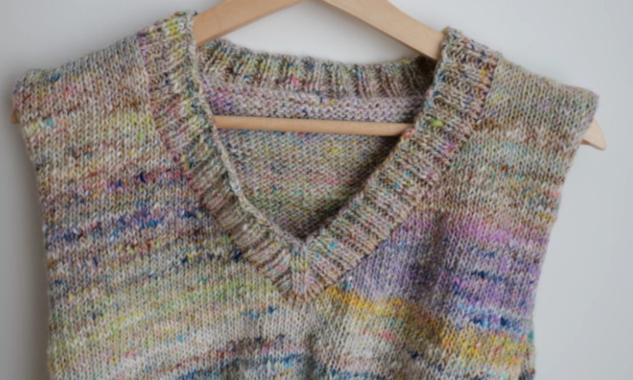 Beige knitted V-neck sweater vest with colorful speckles, hanging on a wooden hanger.