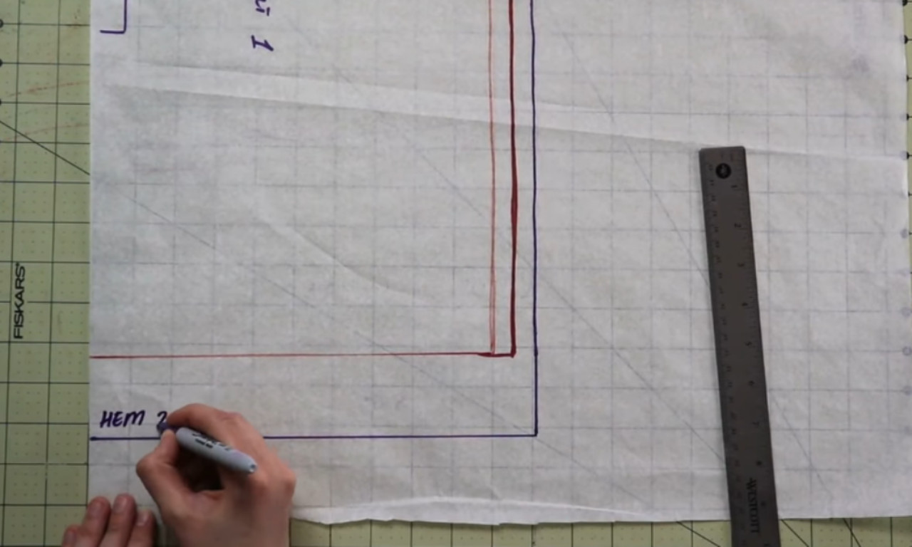 Overhead view of a person labeling a sewing pattern. A left hand holds translucent, gridded pattern paper while the right hand writes “HEM 2” in purple marker along the bottom edge. Red and purple outline lines trace the pattern piece, and a metal ruler rests on a green, gridded cutting mat beneath the paper.