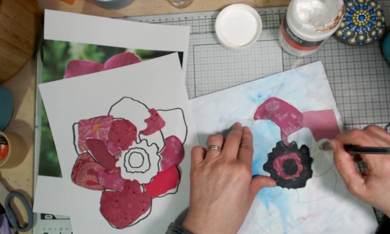 A person is working on a mixed media flower collage. Their left hand is holding a piece of paper with a flower outline, partially filled in with cut pieces of pink and red patterned paper. Their right hand is using a brush to apply adhesive or paint to a similar collage on a separate sheet. Nearby are tools including scissors, a jar of adhesive, and a painted stone. The workspace is organized on a grid cutting mat.