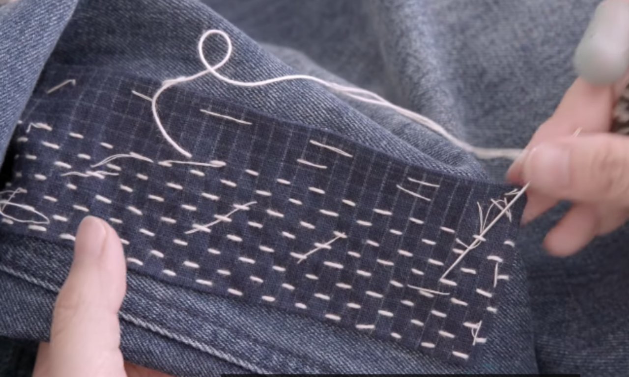 Two hands hold and guide white thread through the fabric of dark blue denim, demonstrating hand-stitching or mending work with visible running stitches along the edge. The focus is on the detailed stitching process against the textured background of the material.