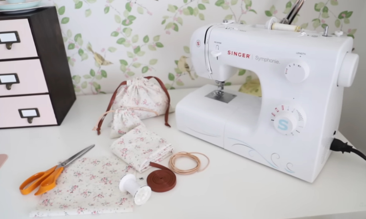 A white Singer sewing machine sits on a white table next to fabric pieces, scissors, and small drawstring bags. The fabric has a floral pattern. A black storage unit is visible in the background, with floral wallpaper behind it.
