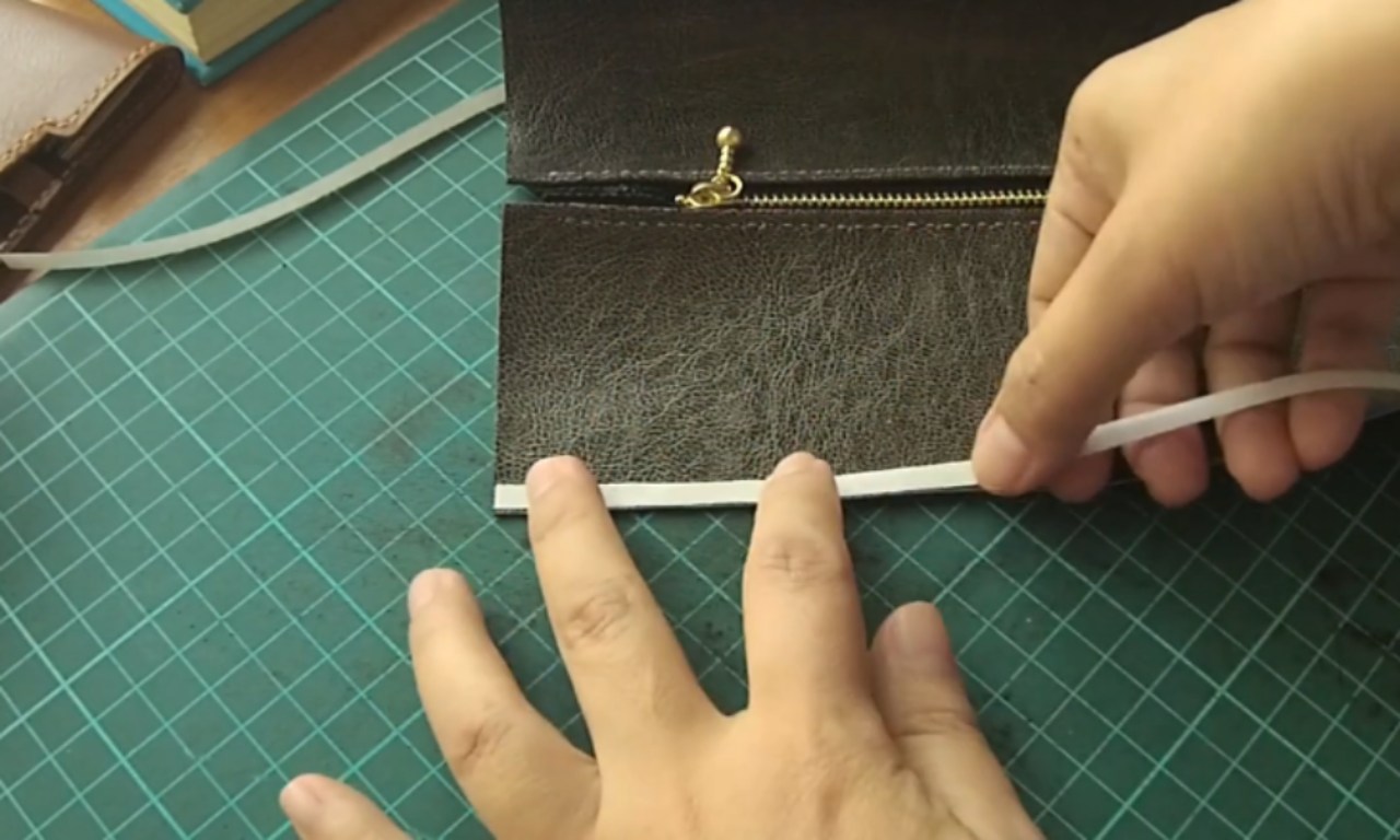 a pair of hands carefully attaching a thin white strip along the bottom edge of a piece of dark brown textured fabric, likely leather or faux leather. A gold zipper and pull are visible at the top of the fabric. The crafting process is taking place on a green self-healing cutting mat with grid lines.