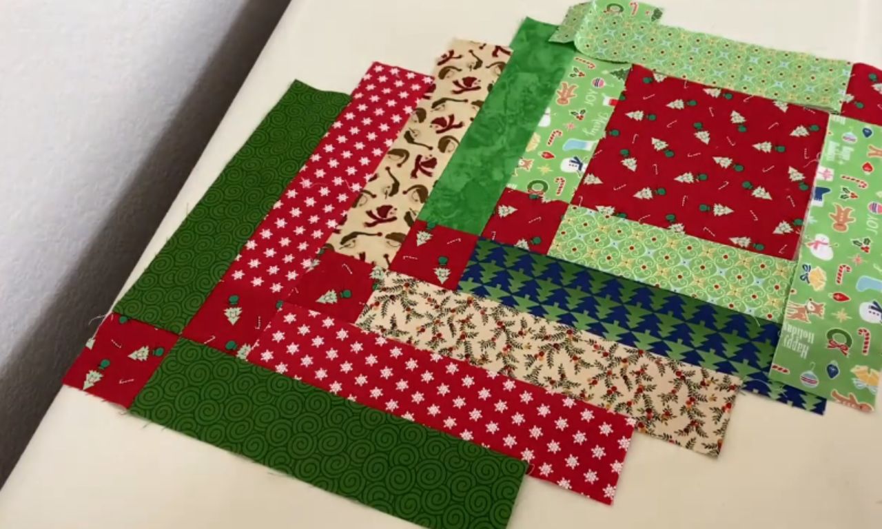 Fabric pieces for a Christmas table runner laid out diagonally, featuring various festive patterns like snowflakes, Santa, Christmas trees, candy canes, holly, and swirls in red, green, cream, and blue.