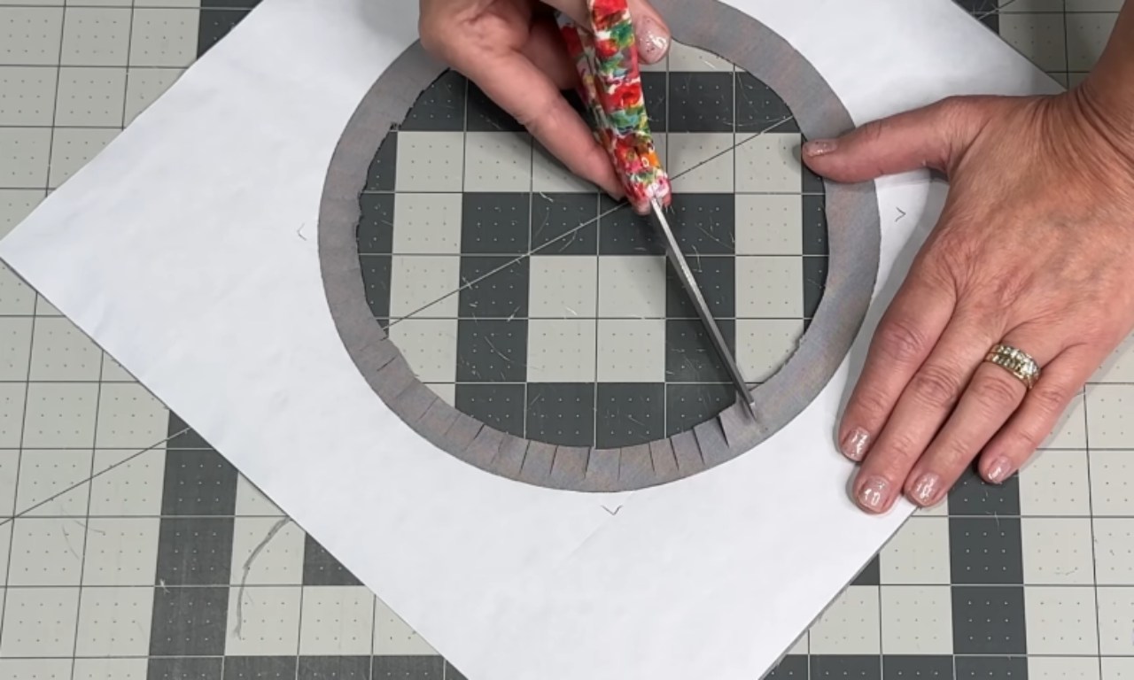 two hands position a floral-handled craft knife (or seam ripper) to clip evenly spaced notches around a grey fabric ring that’s been fused to a sheet of white paper, all resting on a gridded cutting mat.